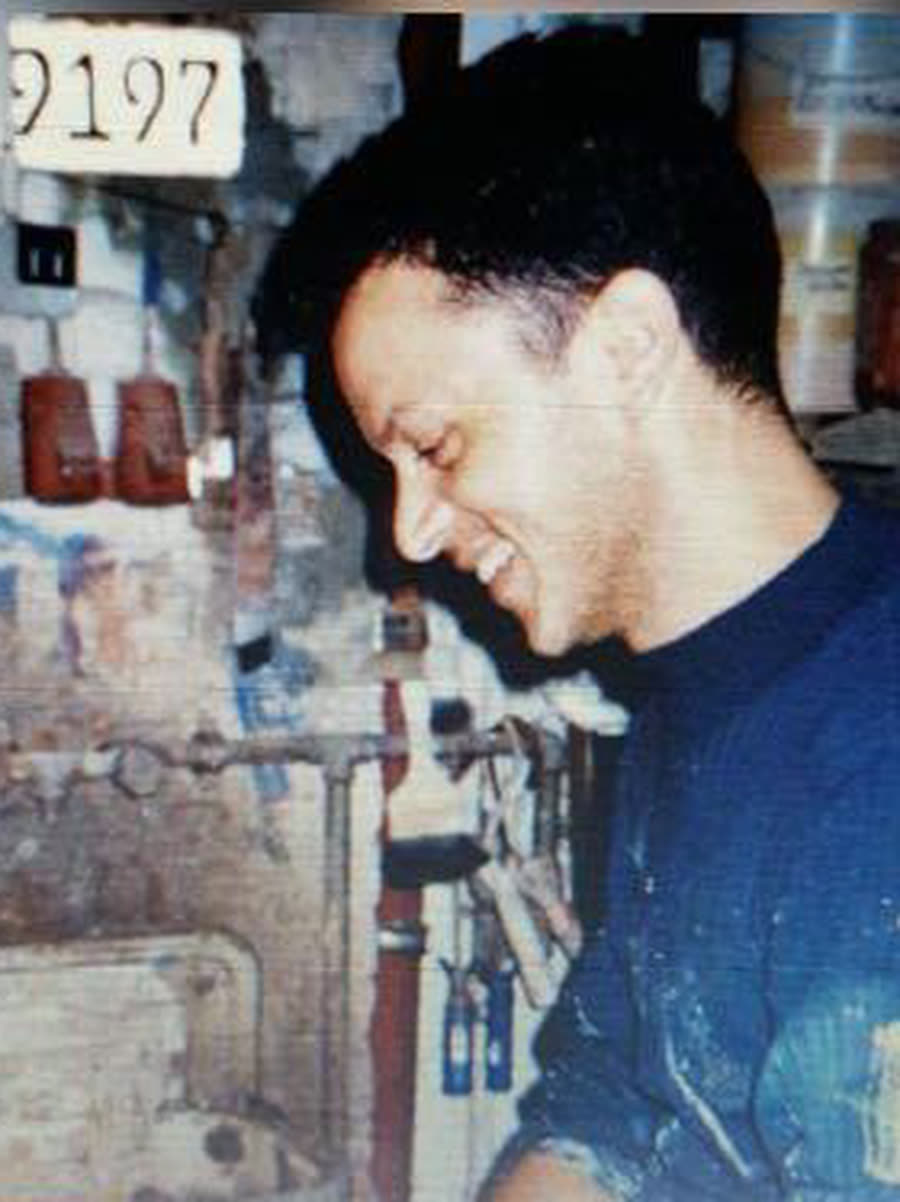 Hafid Azza bei der Arbeit in seiner Werkstatt, lachend, in Arbeitskleidung, umgeben von Werkzeug und Farbdosen